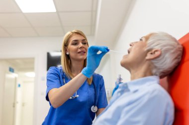 Doktor, yaşlı kadın hastadan boğaz temizleme testi alıyor. Sağlık çalışanı koruyucu iş elbisesinde. Salgın sırasında hastanedeler..