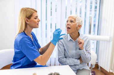 Kıdemli hasta boğazına bakması için ağzını açıyor. Kadın doktor klinikteki hastanın boğazını inceliyor. Otolaryngologist hastanın boğaz ağrısını inceliyor..