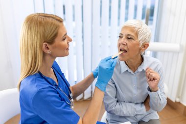 Kıdemli hasta boğazına bakması için ağzını açıyor. Kadın doktor klinikteki hastanın boğazını inceliyor. Otolaryngologist hastanın boğaz ağrısını inceliyor..