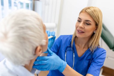 Kıdemli hasta boğazına bakması için ağzını açıyor. Kadın doktor klinikteki hastanın boğazını inceliyor. Otolaryngologist hastanın boğaz ağrısını inceliyor..