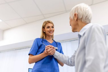 Doktor umut veriyor. Genç kadın hekimin avuçlarında elini tutan yaşlı bayanın gülümsemesini yakından çek. Beyaz önlüklü kadın bakıcı yaşlı insanı cesaretlendiriyor.