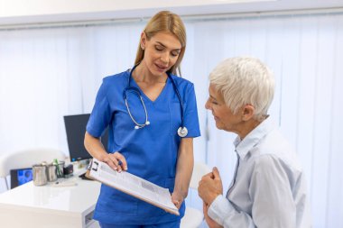 Genç bayan doktor, yaşlı yaşlı bir hastaya danışmanlık yapıyor. Profesyonel doktor üniforması, klinikteki randevuda tıp kağıdı imzalayan olgun bir kadınla konuşuyor..