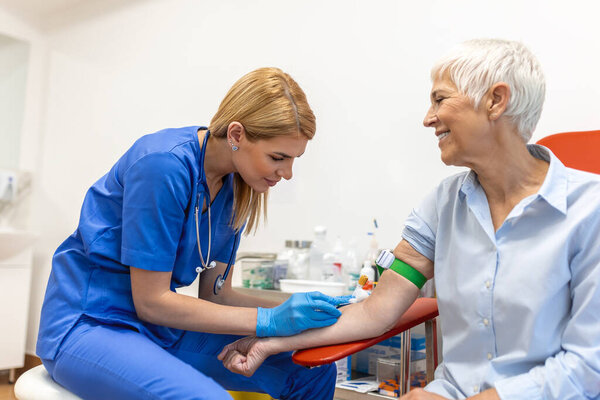 Preparation for blood test with senior woman by female doctor medical uniform on the table in white bright room. Nurse pierces the patient's arm vein with needle blank tube.