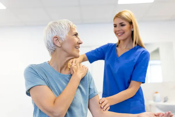快乐的老年妇女和理疗师一起做运动 退休老妇人在疗养期间 在私人教练的帮助下 在诊所展开双臂 图库图片