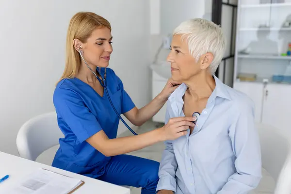 年轻的女医生在临床会面时用听诊器倾听老年病人的心脏胸部 女全科医生用留声机检查女病人 保健概念 图库图片