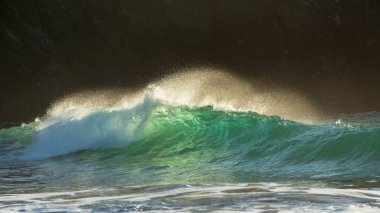 Yeşim turkuaz dalgaların kıyıya çarptığı destansı manzara görüntüsü ve Kynance Cove Cornwall 'daki kayalar, parlayan gündoğumu arka planı ve rüzgardaki su damlacıkları.