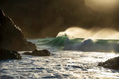 Yeşim turkuaz dalgaların kıyıya çarptığı destansı manzara görüntüsü ve Kynance Cove Cornwall 'daki kayalar, parlayan gündoğumu arka planı ve rüzgardaki su damlacıkları.