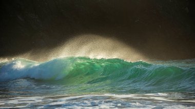 Yeşim turkuaz dalgaların kıyıya çarptığı destansı manzara görüntüsü ve Kynance Cove Cornwall 'daki kayalar, parlayan gündoğumu arka planı ve rüzgardaki su damlacıkları.