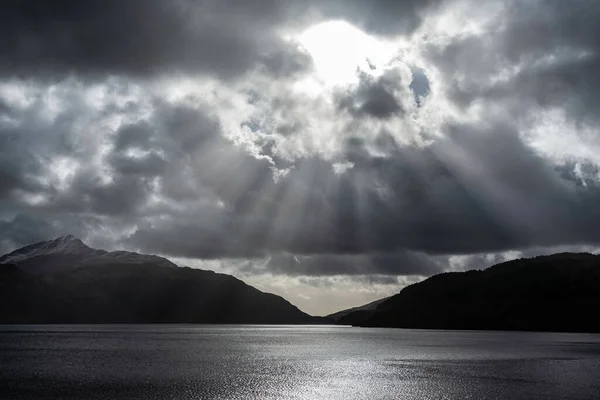 Stunning majestic sunbeams streaming through dramatic clouds onto calm waters of Loch Lomond landscape during Winter sunset
