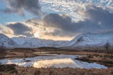 Stunning Winter panorama landscape image of mountain range viewed from Loch Ba in Scottish Highlands with dramatic clouds overhead clipart