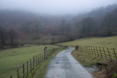 Beautiful Winter landscape image of road around Loughtrigg Tarn on misty morning with calm water and foggy countryside in the background