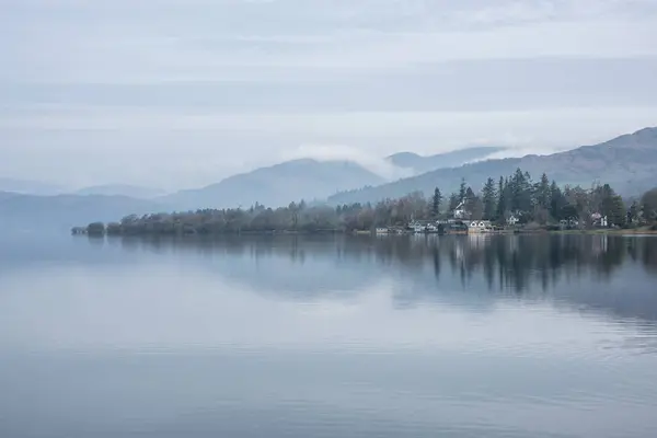 湖と遠くの霧の頂上に船を搭載したウィンダミア湖沿いの霧の朝の美しい平和な風景のイメージ ストック画像