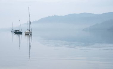 Gölde demirlemiş teknelerle ve uzak sisli tepelerle Windermere Gölü üzerindeki sisli bahar sabahının çarpıcı huzurlu manzarası.
