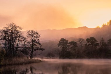 Dramatic atmospheric sunrise landscape at Rydal Water in Lake District during Autumn with moody glow over mist on lake clipart