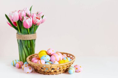 Paskalya ve bahar tatili tebrik kartı konsepti. Sepetinde renkli yumurtalar olan pembe laleler. Yuvaya boyanmış Paskalya yumurtaları ve renkli arka planda laleler, yazı için yer. Boşluğu kopyala.