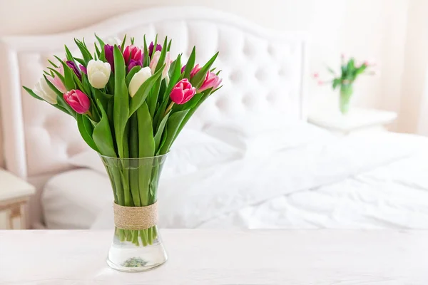 Bedroom in soft light colors. Vase of colorful tulips in light cozy bedroom interior. Home interior with pink tulips in a vase on a light bedroom background.