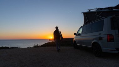 Surferboy sitting near his mini van and looking on the ocean at summer sunset  with a surfboard on her side clipart