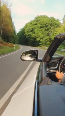 Yetişkin bir adam güneşli bir yaz gününde üstü açık arabasıyla araba sürüyor. Geniş açı takip çekimi yüksek hız hareket bulanıklığı, dikey video