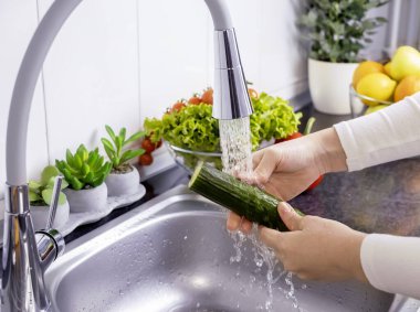 Kadın elleri mutfakta taze salata hazırlamak için salatalık yıkıyor. Sağlıklı çiğ organik yiyecekler, vitaminler, böcek ilacı konseptinden sebzeleri yıkamak..