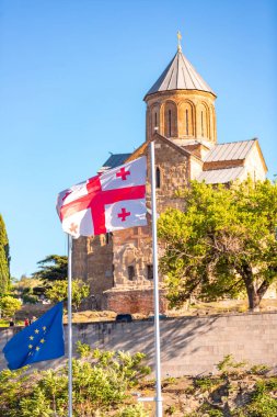 Georgian and EU flags waving in front of the Metekhi church of the Nativity of the Mother of God, located on the left bank of the river Kura, Tbilisi. clipart