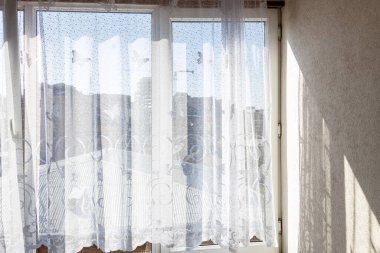 Ev penceresindeki tül perdeler sabah güneşiyle aydınlatılmış ve arka plandaki kentsel evlerin manzarası