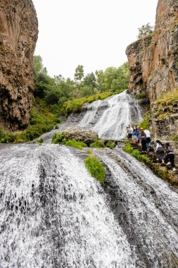 Jermuk, Ermenistan - 3 Ağustos 2024: Turistler yağmurlu yaz günlerinde Arpa nehri vadisindeki Jermuk Şelalesi 'ne tırmanıyor.