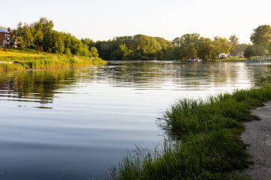 calm surface of Kolomenka river and view of urban beach in Kolomna city at summer sunset clipart