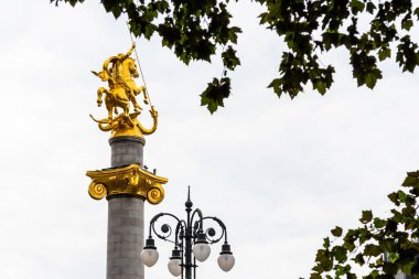 Tbilisi, Georgia - September 22, 2023: green leaves and golden statue St George slaying dragon on top of Liberty Monument on Freedom Square in center of Tbilisi city on cloudy autumn day clipart