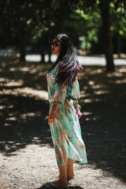 Beyaz elbiseli genç bir kadının dikey fotoğrafı ve elinde bir kitapla parkta pembe bir eşarp.