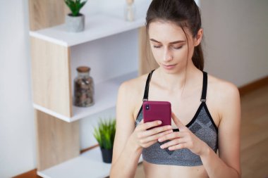 Güzel genç bir kadın akıllı telefonuyla çevrimiçi egzersiz yaparken egzersizleri tekrarlıyor.