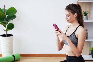 Güzel genç bir kadın akıllı telefonuyla çevrimiçi egzersiz yaparken egzersizleri tekrarlıyor.