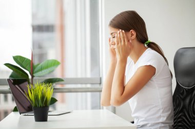 Güzel stresli genç bir ofis çalışanı ofiste acıdan başını tutup oturuyor..