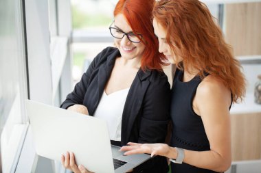 İşyerinde çalışan iki kızıl saçlı iş kadını. Ofisteki bilgisayarlarında bir dizüstü bilgisayar var..