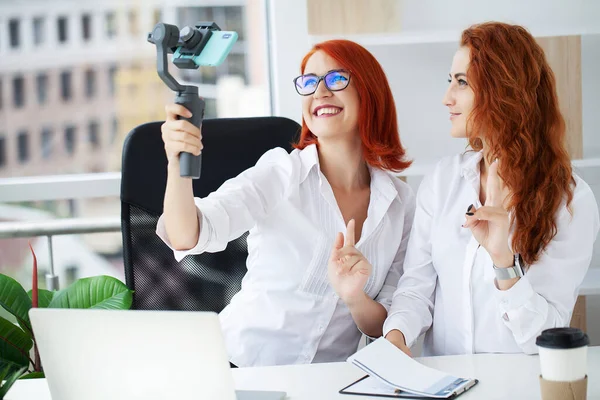 Two red-haired businesswomen in the office recording a video blog.