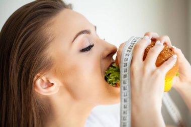 Zararlı hamburgerle beyaz mezura tutan genç bir kadın portresi.