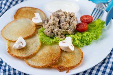 Deruny, mantarlı patatesli gözleme, şampiyonlar, beyaz tabakta ekşi krema. Kirazlı domates, salata. Restoran mavi arka planda servis ediliyor, peçete, çatal, bıçak. Üst görünüm, alanı kopyala