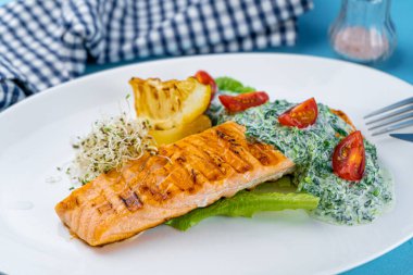 Izgara somon bifteği. Beyaz tabakta bir parça kırmızı balık ve masanın üzerinde mavi bir arka plan. Soslu balık yemi, domates, limon. Restoran menüsü. Kızarmış ızgara