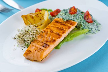 Izgara somon bifteği. Beyaz tabakta bir parça kırmızı balık ve masanın üzerinde mavi bir arka plan. Soslu balık yemi, domates, limon. Restoran menüsü. Kızarmış ızgara