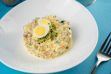 Geleneksel Olivier salatası. Noel ve Yeni Yıl yemekleri. Çatallı beyaz tabak ve mavi arka planda bıçak fotokopi alanı ile. Üst manzara, düz yatış. Mikro yeşil, bıldırcın yumurtası. Bir restoranda hizmet etmek..