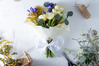 Make bouquet florist workplace. Flower arrangement in a flower shop. Spring bouquet of yellow flowers. Irises, tulips, mimosa and eucalyptus. Tulip bud. Floral background. Purple iris. White table