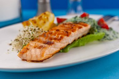 Izgara somon bifteği. Beyaz tabakta bir parça kırmızı balık ve masanın üzerinde mavi bir arka plan. Soslu balık yemi, domates, limon. Restoran menüsü. Kızarmış ızgara
