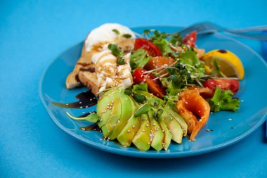 Sağlıklı kahvaltı. Avokado ve somon, haşlanmış yumurta ve tost, salata ve sebze. Tabakta mikroçimler. Yemek kasesi. Mavi tabakta öğle yemeği. Sos ve susam tohumları, limon domatesleri. Üst görünüm.