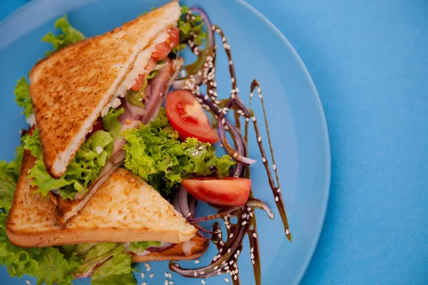 Breakfast sandwich toast with salad. Blue background. Bread, beakon tomatoe on plate top view. Meat sandwich. Sandwiches with ham, cheese, tomatoes, lettuce, and toasted bread. Healthy food.