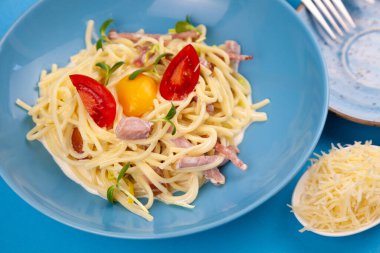 Peynirli ve yumurtalı makarna, pastırma ve domates, mikroyeşil. Mavi tabakta yemek. Ev yapımı carbonara makarnası. Öğle yemeği masanın üst tarafında. Jambonlu İtalyan spagettisi, krema.