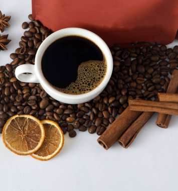 Beyaz bir fincan kahve. Kahve çekirdekleri ve paketleme çantası. Sabah kahvaltısı için canlandırıcı bir içki. Yemek arkaplanı. Uzay afişini kopyala. Tarçın çubukları ve kurutulmuş portakal.