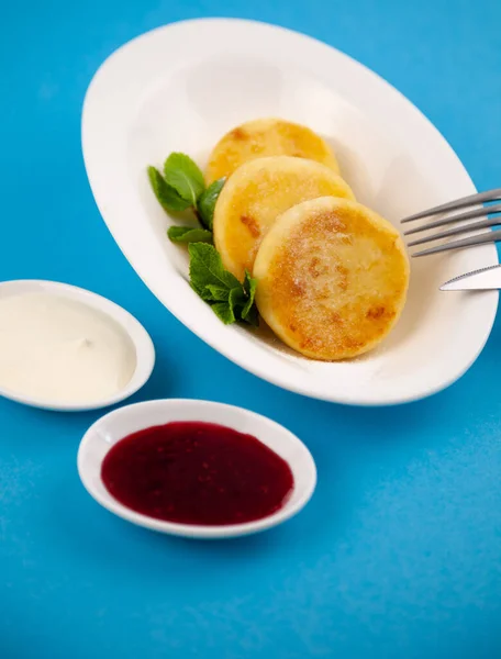 Round cottage cheese pancakes in a white plate on a blue background. Food breakfast. Healthy food. Sour cream sauce and mint. Top view closeup. Dessert meal. Curd cheese pancake ricotta fritters.