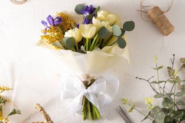 Make bouquet florist workplace. Flower arrangement in a flower shop. Spring bouquet of yellow flowers. Irises, tulips, mimosa and eucalyptus. Tulip bud. Floral background. Purple iris. White table
