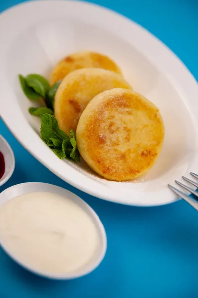 Round cottage cheese pancakes in a white plate on a blue background. Food breakfast. Healthy food. Sour cream sauce and mint. Top view closeup. Dessert meal. Curd cheese pancake ricotta fritters.