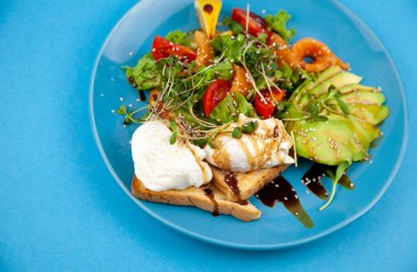 Sağlıklı kahvaltı. Avokado ve somon, haşlanmış yumurta ve tost, salata ve sebze. Tabakta mikroçimler. Yemek kasesi. Mavi tabakta öğle yemeği. Sos ve susam tohumları, limon domatesleri. Üst görünüm.