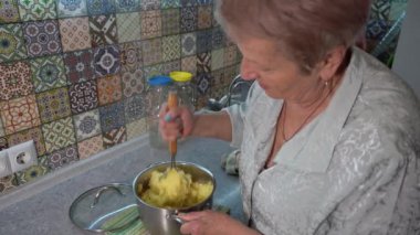 Yaşlı bir kadın mutfakta öğle yemeğinde patates püresi için haşlanmış patates püresi yapar. Eller ve tencere yakın. Yemek masası.
